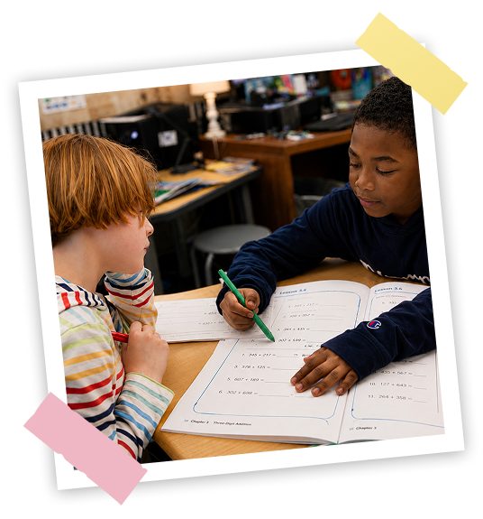 Two children working on a worksheet at a desk in a classroom setting.